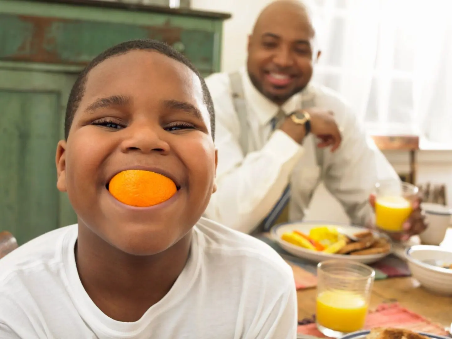 A man and boy sitting at the table eating orange.